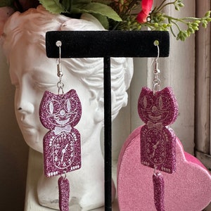 May include: A pair of dangling earrings featuring a glittery purple cat design with a clock face. The earrings have a silver hook and a dangling tail. The cat's face and clock details are white. The earrings are displayed on a black stand.