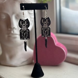 May include: Black cat-shaped earrings with a clock face design. The earrings feature a smiling cat face above a clock, with a dangling tail. The earrings are displayed on a black stand, with a white bust and pink heart in the background.