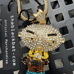 Peut inclure: Un collier en chaîne dorée avec un pendentif en diamant représentant un personnage de dessin animé. Le pendentif porte les mots "Money Maker". La chaîne est de style Cuban link.