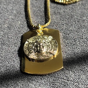 Puede incluir: Un collar con colgante de chapa de perro de color dorado. El colgante presenta un medallón circular con un diseño de rostro detallado, colocado sobre una placa rectangular. La cadena también es de color dorado. El collar se muestra sobre una superficie gris.