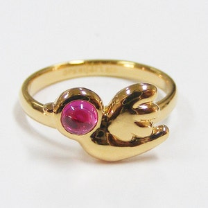 May include: A gold ring featuring a pink gemstone set in the centre. The ring is shaped like a bird with its wings spread.
