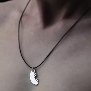 May include: Silver pendant necklace with a black cord. The pendant has a curved shape with a cut-out design. The necklace is worn around the neck.