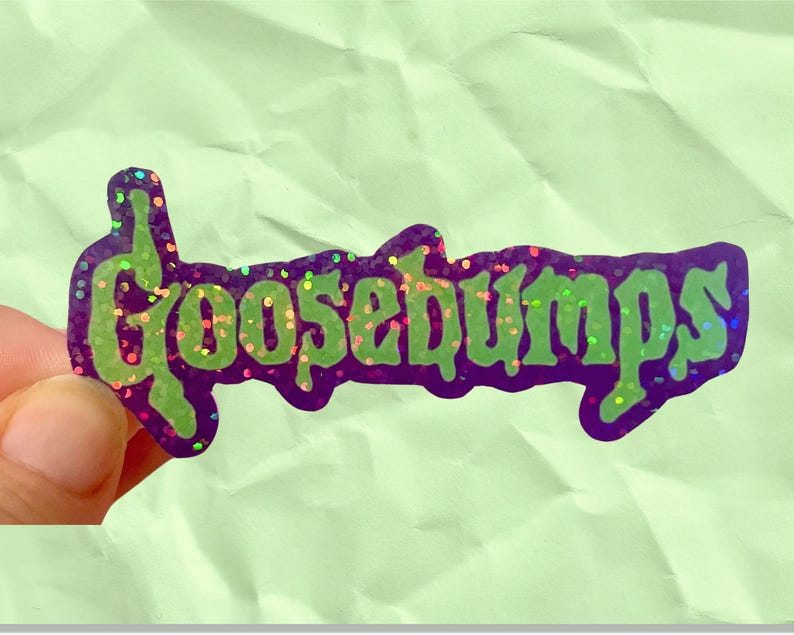 May include: A purple sticker with the word "GOOSEBUMPS" in green, glittery letters. The sticker has a dark purple border and is held against a light green background. The letters have a glittery effect.