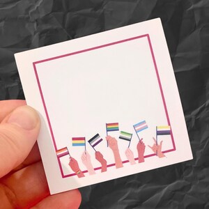 May include: A white square card with a pink border, featuring illustrations of hands holding various LGBTQ+ pride flags. The flags represent different identities and orientations. The card is held against a dark, textured background.