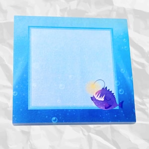 May include: Square sticky note pad with a blue gradient background and a cartoon anglerfish illustration. The anglerfish is purple with a glowing yellow lure. Bubbles are scattered around the edges of the pad.