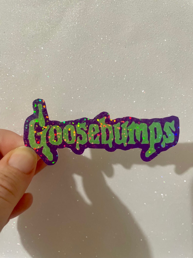 May include: A purple sticker with the word "Goosebumps" in green, glittery letters. The sticker has a dripping font style and a glittery border. The background is white with glitter.