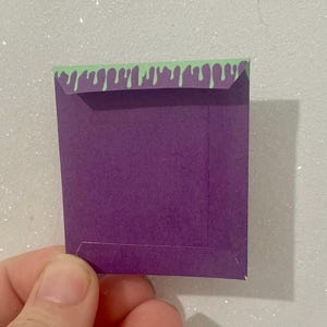 K&ouml;nnte beinhalten: Ein kleiner, quadratischer lila Papierumschlag mit einem gr&uuml;nen, tropfenden Schleimdesign am oberen Rand. Der Umschlag wird in einer Hand vor einem glitzernden, wei&szlig;en Hintergrund gehalten. Der Umschlag scheint handgefertigt zu sein.