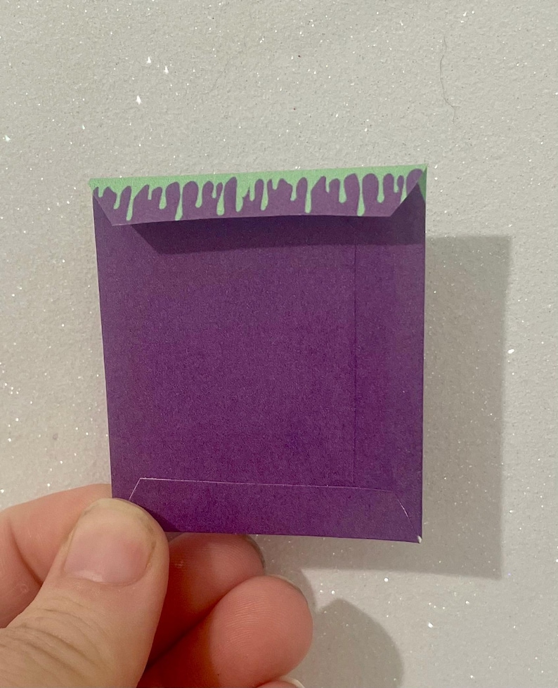 K&ouml;nnte beinhalten: Ein kleiner, quadratischer lila Papierumschlag mit einem gr&uuml;nen, tropfenden Schleimdesign am oberen Rand. Der Umschlag wird in einer Hand vor einem glitzernden, wei&szlig;en Hintergrund gehalten. Der Umschlag scheint handgefertigt zu sein.
