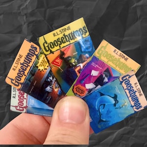 K&ouml;nnte beinhalten: Eine Hand h&auml;lt einen F&auml;cher aus Miniatur-Goosebumps-B&uuml;chern. Die Buchcover zeigen Illustrationen in verschiedenen Farben, darunter Orange, Blau und Gr&uuml;n, wobei der Serientitel deutlich sichtbar ist. Die B&uuml;cher stammen aus der R.L. Stine-Reihe.