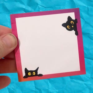May include: A square, white sticky note with a pink and purple border. Two black cat illustrations with yellow eyes are on the corners. The note is held by a hand against a blue background.
