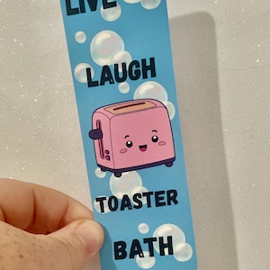 May include: A blue bookmark with white bubbles and the text "Live Laugh Toaster Bath". A pink cartoon toaster with a smiling face is in the center of the bookmark.