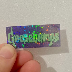 K&ouml;nnte beinhalten: Ein rechteckiger Aufkleber mit dem Wort "Goosebumps" in gr&uuml;ner, tropfender Schrift. Der Aufkleber hat einen lila Hintergrund mit schillerndem Glitzer. Der Aufkleber wird von den Fingern einer Person gehalten.