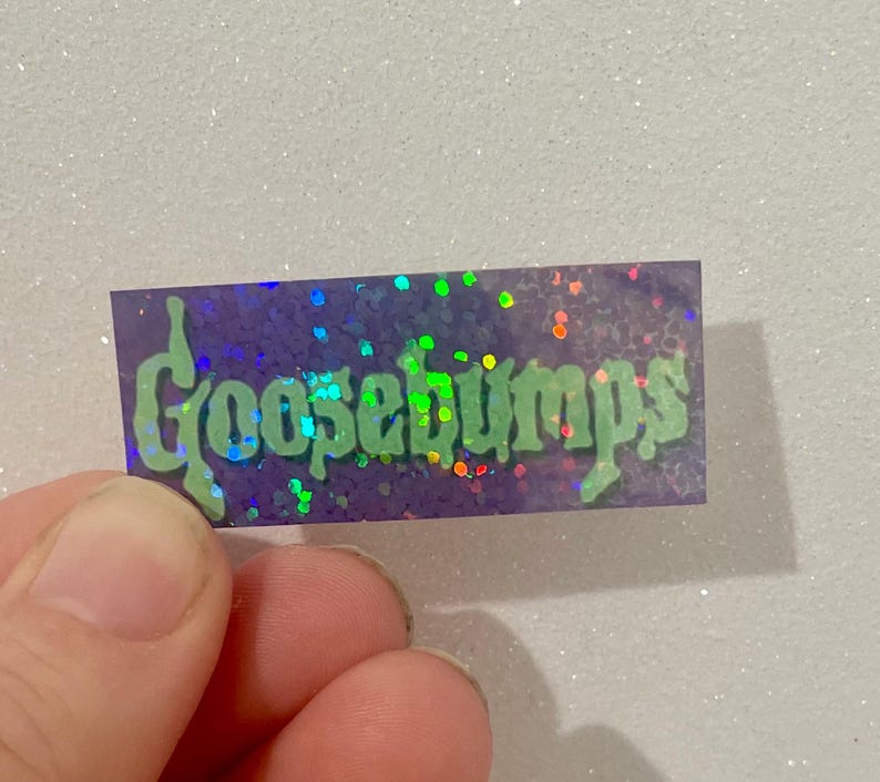 K&ouml;nnte beinhalten: Ein rechteckiger Aufkleber mit dem Wort "Goosebumps" in gr&uuml;ner, tropfender Schrift. Der Aufkleber hat einen lila Hintergrund mit schillerndem Glitzer. Der Aufkleber wird von den Fingern einer Person gehalten.