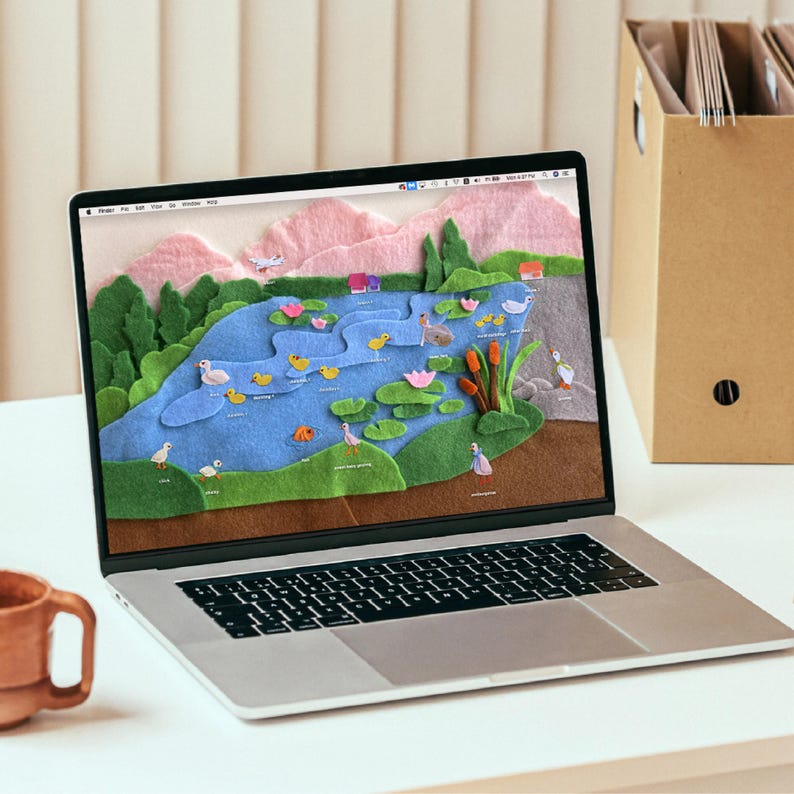 May include: A laptop displays a felt art scene of a pond with ducks, fish, and lily pads. The scene includes pink mountains, green trees, and a blue pond. A brown mug sits to the left of the laptop.