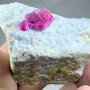 May include: A rough-cut mineral specimen featuring a vibrant, hot pink ruby crystal cluster atop a white matrix. The ruby crystals display a textured surface, contrasting with the matrix's smoother appearance. The base of the specimen has a layered, earthy tone.