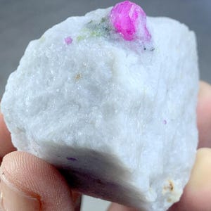 May include: A rough, white mineral specimen with a vibrant pink crystal formation on top. The crystal has a faceted appearance, and there are small flecks of green and black minerals present.