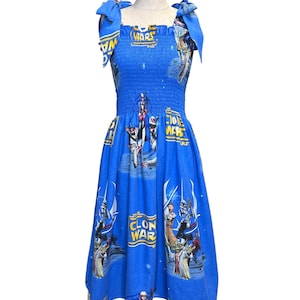May include: A blue midi dress featuring a smocked bodice and tie-shoulder straps. The fabric displays a Star Wars The Clone Wars print with various characters, including Yoda and clone troopers, alongside the yellow and blue logo text.