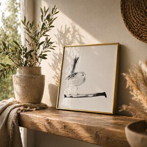 May include: A square framed wall art piece featuring a black ink illustration of a small bird perched on a branch. The artwork is set within a thin gold-colored metal frame, showcasing a minimalist bird drawing on a plain white background.