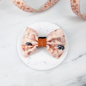 Puede incluir: Una pajarita color melocotón con un estampado de animales del bosque, incluyendo ardillas, tejones y búhos. El lazo tiene un centro marrón y está sobre una superficie de mármol blanco. Una cinta a juego está en el fondo.