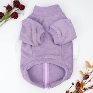 Puede incluir: Una chaqueta para perros de forro polar lavanda con cuello alto y mangas largas. La chaqueta tiene una cremallera de largo completo en la parte delantera y una raya lavanda en contraste. La chaqueta está diseñada para mascotas.