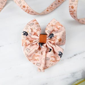 May include: A large, pink fabric bow with a woodland animal pattern, including deer, owls, and badgers. The bow has a brown center knot and is set against a white background. A matching ribbon is visible in the background.