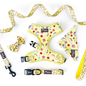 Könnte beinhalten: Ein gelbes Hundegeschirr, eine Leine, ein Halsband und ein Bandana mit einem Einhorn-, Cupcake- und Regenbogenmuster. Die Accessoires sind alle mit dem Wort "Piggie" beschriftet.