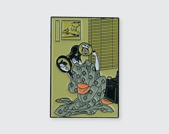 May include: A green and gold enamel pin depicting a woman in a traditional Japanese kimono holding a mirror and looking at her reflection. The background is a yellow room with a window and blinds.