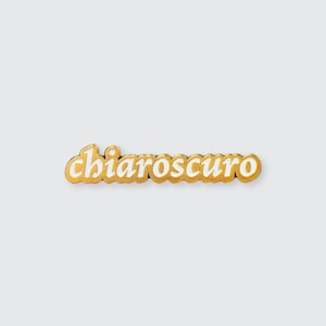 May include: Gold-coloured pin with the word "chiaroscuro" written in white letters.