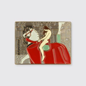 May include: A gold-toned enamel pin featuring a woman riding a white horse. The woman is nude and has long brown hair. The horse is wearing a red and green saddle. The background is a cityscape with buildings and trees.