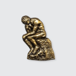 May include: A gold-colored sculpture of a man sitting and thinking, known as "The Thinker".