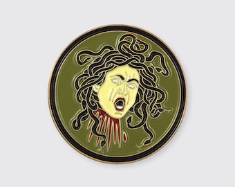 May include: A round, green enamel pin with a black outline. The pin features a detailed illustration of Medusa's head with snakes for hair. Her face is yellow with red lips and a red tongue.