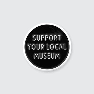 Op de afbeelding: Zwarte en witte ronde pin met de tekst "Support Your Local Museum" in witte letters.