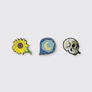 May include: Three enamel pins featuring famous artwork. The first pin is a yellow sunflower with green leaves. The second pin is a blue and gold swirling night sky with a crescent moon. The third pin is a white skull with a green jaw and a cigarette in its mouth.