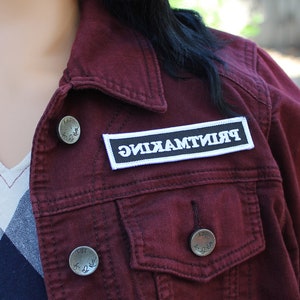 May include: A maroon denim jacket with a white patch that says "PRINTMAKING" on it. The jacket has two silver buttons with the words "KUT" and "For Life" on them.