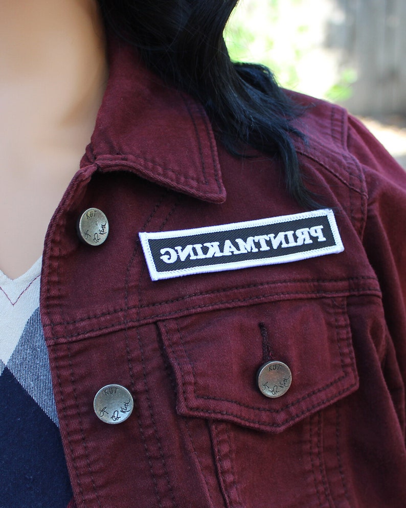 May include: A maroon denim jacket with a white patch that says "PRINTMAKING" on it. The jacket has two silver buttons with the words "KUT" and "For Life" on them.
