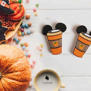 May include: Two orange and brown Halloween-themed coffee cups with Mickey Mouse ears. The cups are on a white wooden surface with a pumpkin and Halloween cookies in the background.