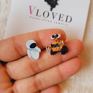May include: A pair of handcrafted jewelry earrings featuring the characters Wall-E and Eve from the animated film. The Wall-E earring is orange and black, while the Eve earring is white and blue. The earrings are displayed on a white card with the text "Vloved Handcrafted Jewelry".