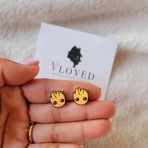 May include: A pair of yellow and green cartoon character stud earrings. The character has a large head with a small body and large eyes. The earrings are on a white background.