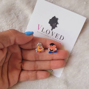 May include: A pair of earrings featuring Mr. and Mrs. Potato Head from the Toy Story movie. The earrings are made of polymer clay and are painted in bright colors. The earrings are on a white background with a business card that says "VLoved Handmade Jewelry".