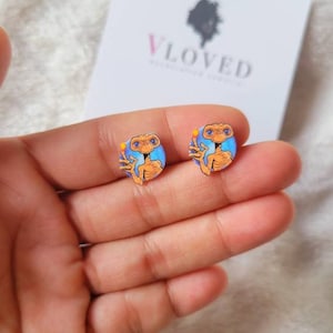 May include: A pair of stud earrings featuring a cartoon image of the alien character E.T. from the movie E.T. the Extra-Terrestrial. The earrings are round and have a blue background with a yellow glow around E.T.'s finger.