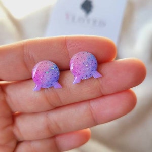 May include: A pair of pink and purple glitter stud earrings shaped like a sphere with a small, flat base. The earrings have a subtle ombre effect, transitioning from pink to purple.