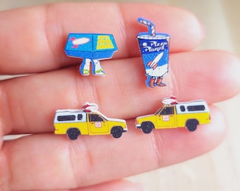 Op de afbeelding: Een set van vier kleurrijke, cartoonachtige, houten oorbellen. De oorbellen hebben een blauwe doos met benen, een blauwe beker met een rietje en twee gele pick-up trucks. De trucks hebben een wit dak en een rood lampje bovenop.