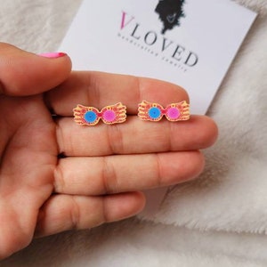 May include: A pair of gold-tone earrings with a pink and blue enamel design. The earrings are shaped like a pair of glasses with a lightning bolt on each side. The earrings are on a white fuzzy surface.