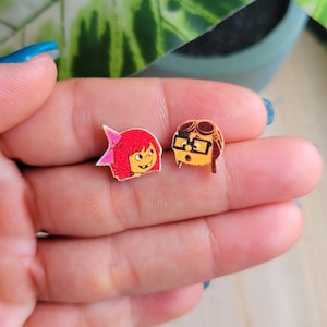 May include: A pair of stud earrings featuring cartoon characters. The left earring is a red-haired girl with a pink bow. The right earring is a brown-haired boy wearing glasses.