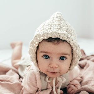 Könnte beinhalten: Ein Baby trägt eine cremefarbene Häkelmütze mit Schleife und liegt auf einer rosa Decke.