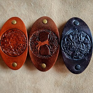 May include: Three oval-shaped leather keychains in shades of brown and black. Each keychain features a detailed embossed design and a brass rivet. The designs include a tree, a deer, and an abstract pattern.