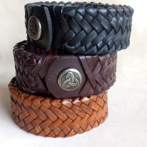 K&ouml;nnte beinhalten: Drei geflochtene Lederarmb&auml;nder mit Metallakzenten. Das obere Armband ist schwarz, das mittlere Armband ist braun und das untere Armband ist beige.