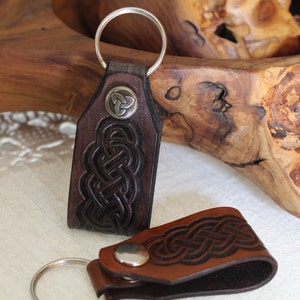 Op de afbeelding: Twee leren sleutelhangers met Keltische knoopontwerpen. De sleutelhanger aan de linkerkant heeft een zilveren ring en een zilveren Keltische knoopbedel. De sleutelhanger aan de rechterkant heeft een bruin leren bandje met een zilveren ring.