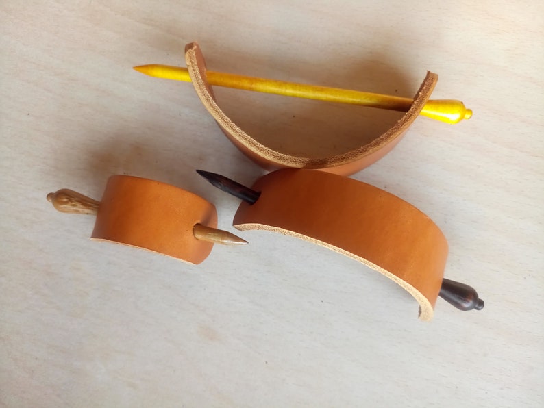 May include: Three leather hair clips with wooden dowels. The clips are brown leather and the dowels are natural wood, black, and yellow.
