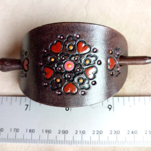 Może przedstawiać: Brązowa sk&oacute;rzana spinka do włos&oacute;w z ozdobnym wzorem w postaci pomarańczowych i r&oacute;żowych serc i kwiat&oacute;w. Spinka ma około 18 cm długości.